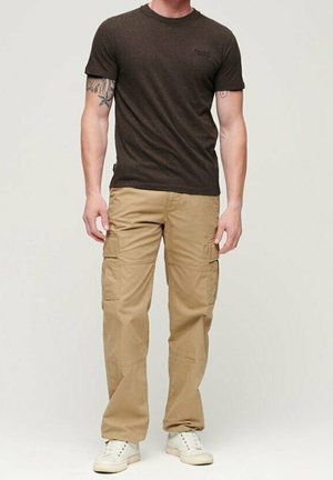 Homme portant un t-shirt marron foncé à manches courtes, un pantalon cargo beige, des baskets blanches, avec des tatouages visibles sur l'avant-bras gauche et le biceps droit.