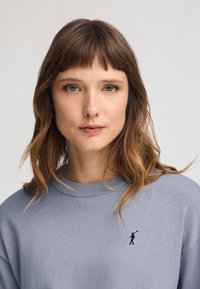 Pull gris avec un col côtelé et un petit personnage brodé sur la poitrine. Texture douce, coupe décontractée et manches longues.