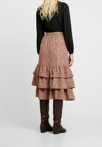 Jupe maxi avec un motif floral marron, ornée de couches volantes, avec une taille élastique, associée à des bottes marron texturées arrivant au genou. Haut à manches longues noir.