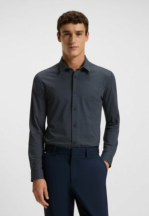 Dunkelblaue Button-Up-Hemd mit weißem geometrischen Muster, langen Ärmeln und Kentkragen; kombiniert mit dunkelgrauen maßgeschneiderten Hosen. Glatte Stofftextur.