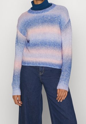 Femme portant un pull tricoté dégradé bleu pastel et rose sur un col roulé bleu marine, associé à un jean taille haute à jambes larges bleu foncé.