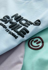 Lichtblauw katoenen overhemd met zwarte verhoogde tekst "NICE SMILE" en "keep it going," gecombineerd met mintgroene en lavendelkleurige stoffen delen en een logodetail.