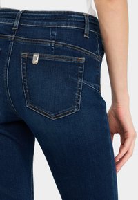 Gros plan sur une personne portant un jean en denim bleu foncé avec une poche arrière visible et une main reposant le long de la cuisse.