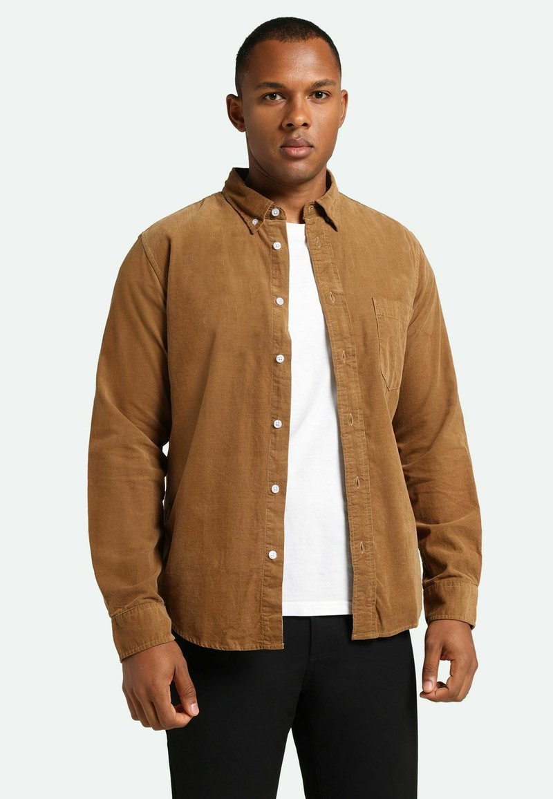 Camisa de pana marrón con botones blancos y un bolsillo. Llevada sobre una camiseta blanca, la camisa presenta un cuello y un ajuste relajado.