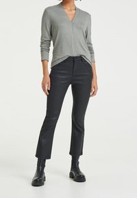 Kvinna klädd i en grå långärmad V-ringad topp instoppad i svarta panoramadräkter och svarta ankelstövlar, stående mot en enkel bakgrund.