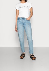Bílá tričko s potiskem "CALVIN KLEIN JEANS", světle modré džíny s vysokým pasem a černé sandály se řemínky, umístěné na šedé podlaze.