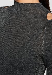 Haut en maille noire pailletée avec col montant et découpe à l’épaule, montrant un tissu texturé ainsi qu’un cou et une épaule partiels.