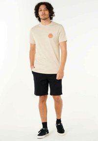 Beige t-shirt med korte ærmer og en rund orange grafik på venstre bryst, parret med sorte shorts og sorte sneakers. Enkel design.