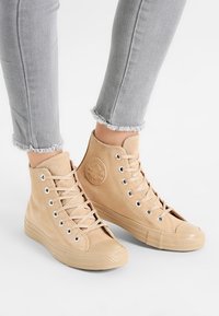 Beige semišové vysoké tenisky s kulatou špičkou, gumovou podrážkou, kovovými očky a kulatým logem na boku. Texturovaný materiál.