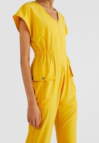 Gul overall i mjuk, lättvikts tyg. Har V-ringning, korta ärmar, elastisk midja och två framfickor med tryckknappar.