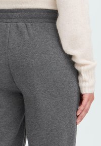 Detailný záber na ruku osoby odpočívajúcu pri jej boku, oblečenú v krémovom svetri a šedých elastických nohaviciach.