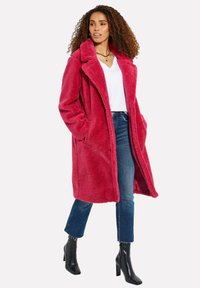 Fuzzy rosa kappa med slagkrage, sidofickor och lång skärning, lager på en vit V-ringad skjorta och tillsammans med blå jeans och svarta stilettklackar.