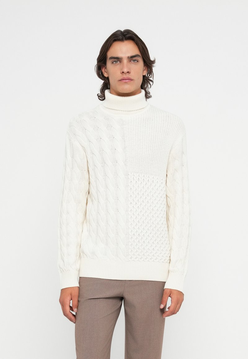 Mann, der einen cremefarbenen grob gestrickten Rollkragenpullover und taupefarbeneHosen trägt, vor einem schlichten weißen Hintergrund steht und nach vorne schaut.