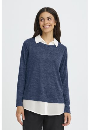 Femme souriante, portant un pull à manches longues bleu marine par-dessus une chemise blanche à col, avec un pantalon noir, debout devant un fond clair uni.