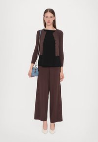 Cardigan marron côtelé sur un haut noir, associé à un pantalon large marron. Porte un sac à main bleu et des chaussures à talons blanches.