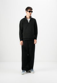Sudadera de forro polar negro con cremallera de un cuarto y bolsillo frontal, combinada con pantalones negros holgados y zapatillas azules, llevada con gafas de sol.