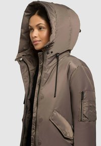Braune gesteppte Pufferjacke mit großer Kapuze, Reißverschluss und Druckknopfverschlüssen, seitlichen und Armstaschen sowie verstellbaren Kordelzügen.
