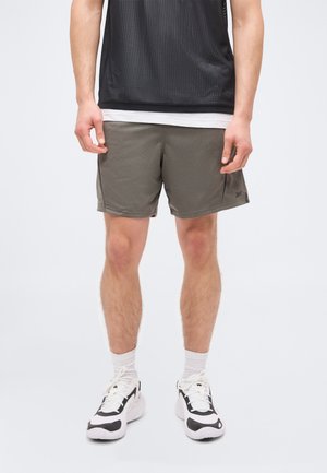 Homem vestido com calções desportivos cinzentos em malha, meias brancas e ténis pretos e brancos, em pé contra um fundo liso.