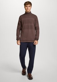 Man die naar voren loopt in een bruine coltrui, marineblauwe rechte broek en donkerbruine laarzen tegen een effen achtergrond.