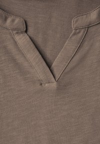 Ruskea kangastoppi, jossa on V-pääntie ja vahvistettu nappilista. Kankaan pinta on pehmeä, siinä on hillitty heather-kuvio ja näkyvät ompeluyksityiskohdat.