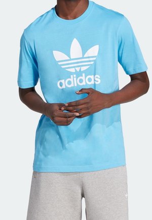 Homme portant un T-shirt Adidas bleu clair avec logo blanc et short gris, mains jointes au niveau de la taille.