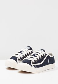 Navy blue canvas sneakers with white rubber sole, rounded toe, white laces, and metal eyelets; featuring logo accents on sides and tongue.