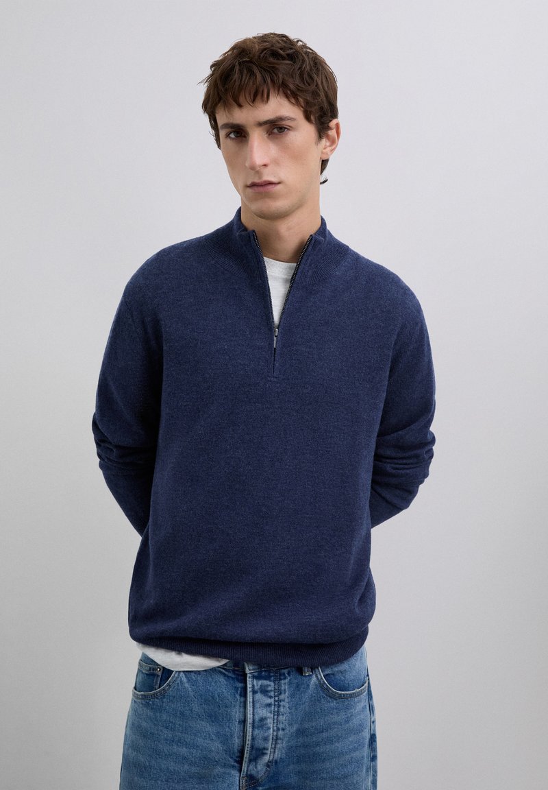 Young man with short brown hair wearing a navy blue quarter-zip sweater over a white shirt and blue jeans, standing against a plain gray background.
