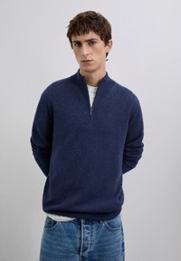 Jonge man met kort bruin haar, gekleed in een marineblauwe trui met een kwart rits over een wit shirt en blauwe jeans, staande tegen een effen grijze achtergrond.