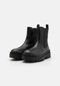 Bottes chelsea en cuir noir avec des panneaux élastiques sur les côtés, semelle en caoutchouc épaisse, bout arrondi et tirant à l'arrière. Texture lisse, design minimal.