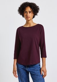 Burgunderfarbenes Oberteil mit drei Viertelärmeln und weitem Ausschnitt, aus weichem Stoff gefertigt. Kombiniert mit blauen Jeans im Denim-Stil. Schlichte, lässige Passform.