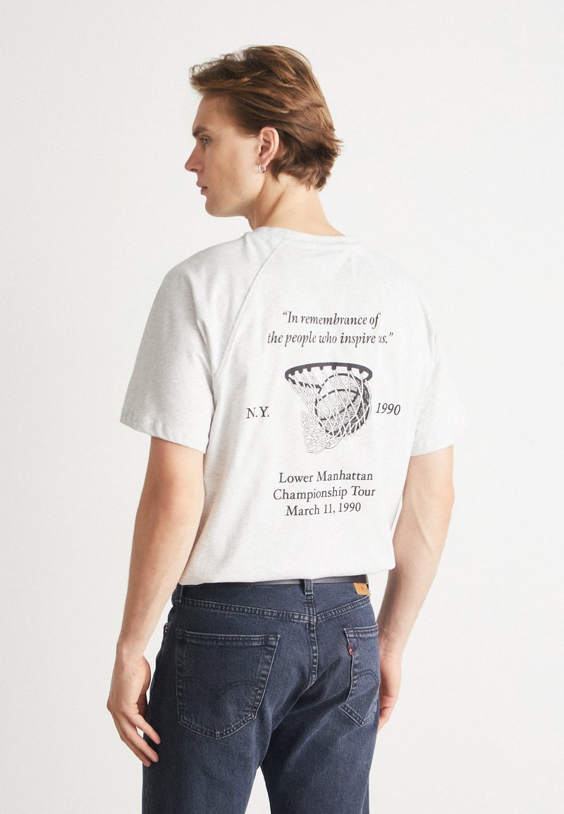 Tricou gri din bumbac cu guler rotund, având un grafic de baschet și text pe spate. Purtați-l cu blugi negri.