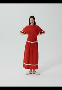 Femme debout, les mains sur les hanches, portant un haut rouge à boutons et une longue jupe assortie avec un bord blanc, associée à des chaussures plates blanches.