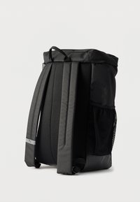 Μαύρο σακίδιο με γεμισμένους ιμάντες, πίσω πάνελ από δίχτυ, πλαϊνή τσέπη από δίχτυ, φερμουάρ στο επάνω μέρος και μια μικρή θηλιά στο χέρι, σε λευκό φόντο.