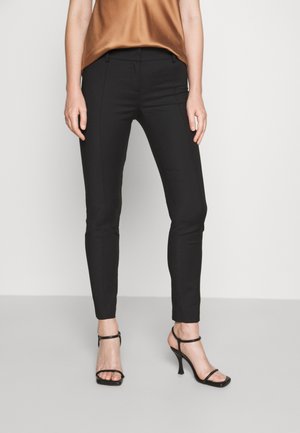 Femme portant un pantalon slim noir longueur cheville, un haut marron sans manches et des sandales à talons noirs, debout devant un fond uni.