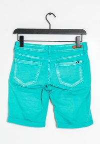Pantalones cortos de mezclilla turquesa colgados en una percha negra. Las características incluyen puños enrollados, dos bolsillos traseros y un acento de cuero cerca de la cintura.