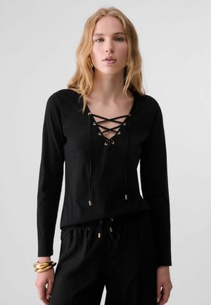Giovane donna con lunghi capelli biondi che indossa una maglia nera a maniche lunghe con scollo allacciato e pantaloni neri coordinati, accessoriata con braccialetti dorati.