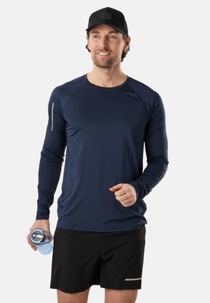 Mężczyzna w czarnej czapce, granatowej koszuli z długim rękawem, czarnych szortach, trzymający butelkę wody, uśmiechający się, stojący na białym tle.