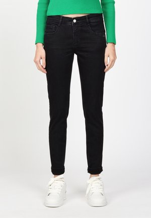 Femme portant un jean noir ajusté, un haut à manches longues côtelé vert, et des baskets blanches, debout devant un fond clair uni.