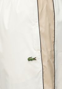 Λευκά σορτς με λεία υφή, που διαθέτουν ένα ανοιχτό μπεζ πλαϊνό πάνελ και σκούρο φινίρισμα. Περιλαμβάνει ένα μικρό κεντημένο λογότυπο κροκοδείλου σε πράσινο χρώμα.