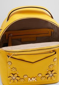 Mochila de cuero amarilla con cierre de cremallera, bolsillos interiores, adornos florales y herrajes en tono dorado.