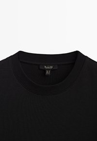 T-shirt noir col rond avec col côtelé et étiquette de marque visible cousue à l'intérieur de l'encolure indiquant la taille petit.