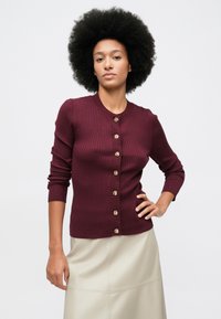 Mulher com cabelo afro natural, vestindo um cardigan justo em ribana bordeaux com botões dourados e uma saia creme, em pé com uma mão na anca.