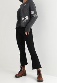 Osoba ubrana w ciemnoszary sweter z haftowanym florystycznym wzorem, czarne spodni cropped flare oraz brązowe sznurowane botki, stojąca na białym tle.