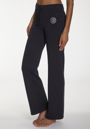 Woman wearing black wide-leg sweatpants with drawstring waist and circular logo on left hip, standing barefoot on white floor.