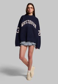 Maglione oversize navy in maglia con scritte bianche "WRSTBHVR" e numero "23". Abbinato a una gonna mini in denim e sneakers crema.