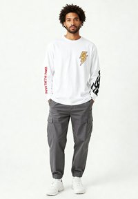 Man met krullend haar, gekleed in een wit shirt met lange mouwen met afbeeldingen van bliksemschichten en vlammen, grijze cargobroek en witte sneakers.