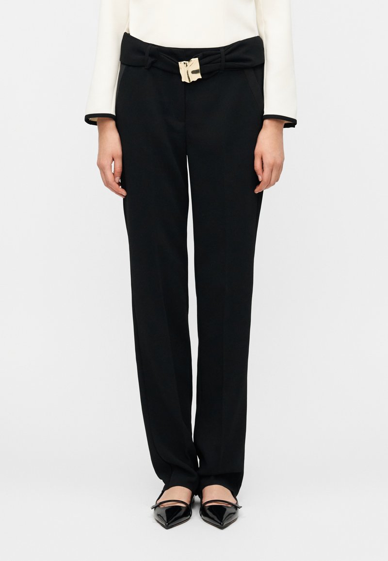 Μαύρο ραμμένο παντελόνι με ομαλή υφή, που διαθέτει ζώνη με φιόγκο και χρυσές μεταλλικές λεπτομέρειες, συνδυασμένο με μαύρα μυτερά φλατ παπούτσια.