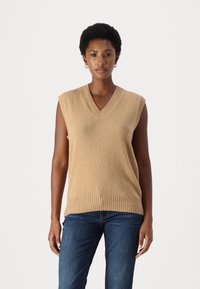 Beige ärmelloser Strickpullover mit V-Ausschnitt, geripptem Saum und Ausschnitt, kombiniert mit dunkelblauen Jeans. Weiche Textur und lässige Passform.