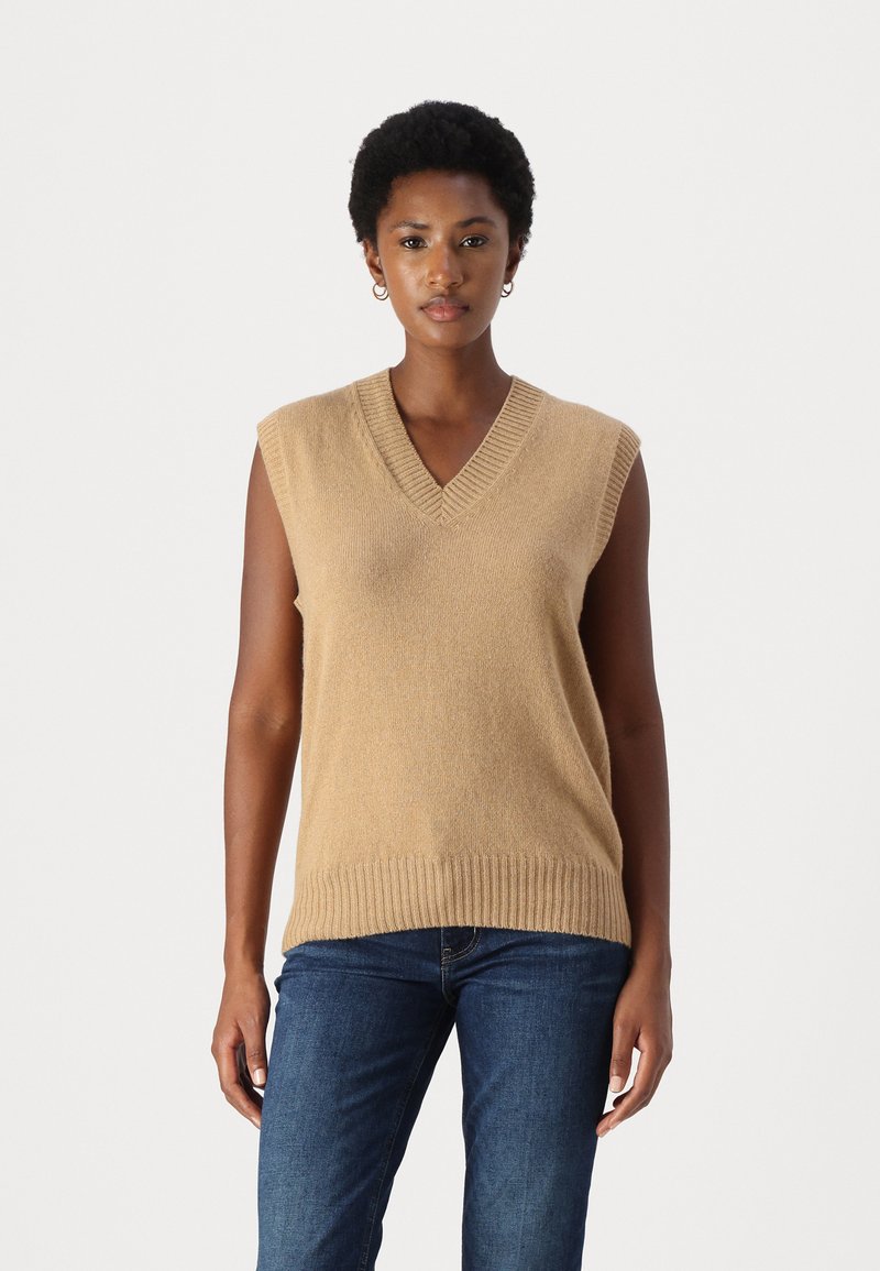 Beige ärmelloser Strickpullover mit V-Ausschnitt, geripptem Saum und Ausschnitt, kombiniert mit dunkelblauen Jeans. Weiche Textur und lässige Passform.
