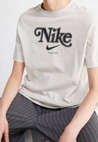 Světle béžové bavlněné tričko s černým logem Nike a textem "Od roku 1972". Kulatý výstřih, krátké rukávy, hladká textura, uvolněný střih.